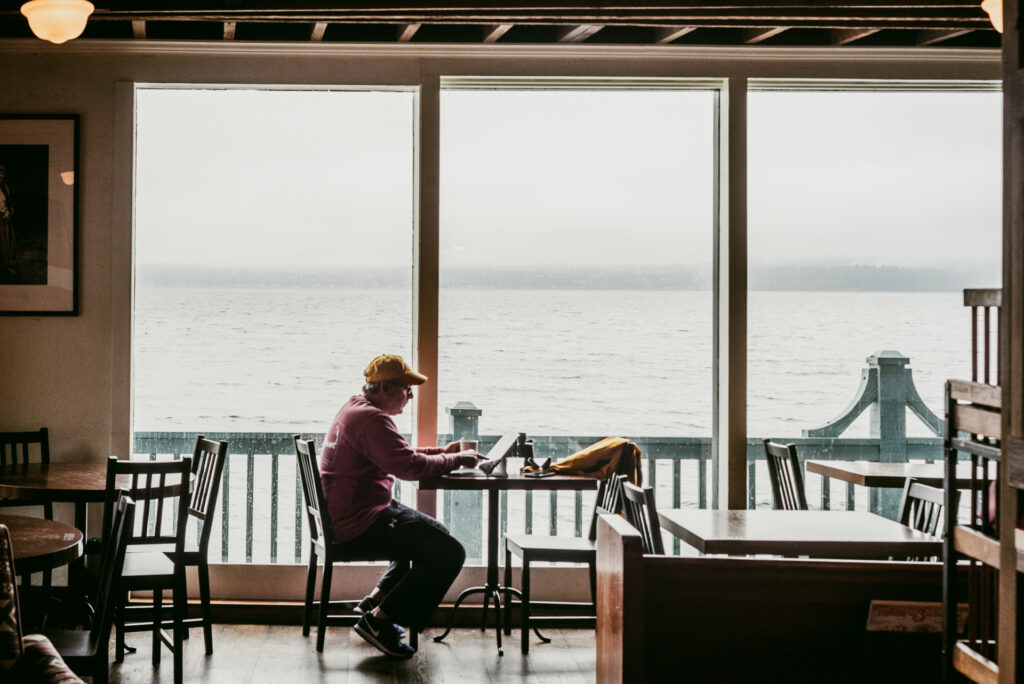 Un homme assis dans un café regarde une tablette posée sur une table, face à une baie vitrée donnant sur l'eau brumeuse.