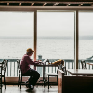 Un homme assis dans un café regarde une tablette posée sur une table, face à une baie vitrée donnant sur l'eau brumeuse.