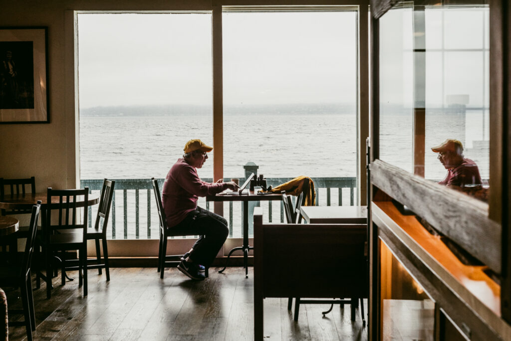 Un homme avec une casquette jaune est assis à une table de café, un ordinateur portable devant lui, face à une vue sur l'eau.