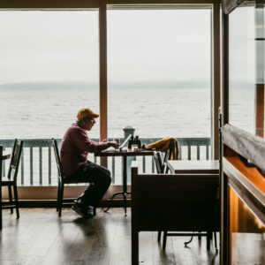 Un homme avec une casquette jaune est assis à une table de café, un ordinateur portable devant lui, face à une vue sur l'eau.