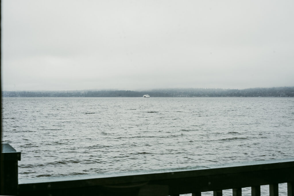 Vue depuis une jetée en bois sur un ferry naviguant au loin sur une eau calme, sous un ciel brumeux et gris.