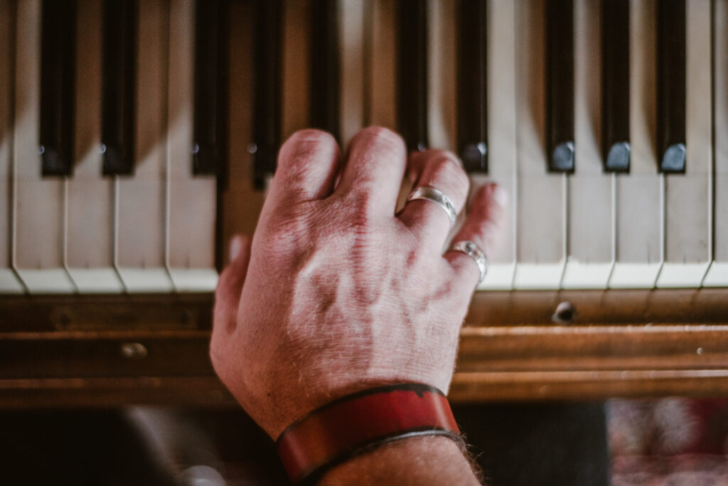 Une main portant des bagues en argent et un bracelet en cuir rouge repose sur les touches d'un piano ancien.