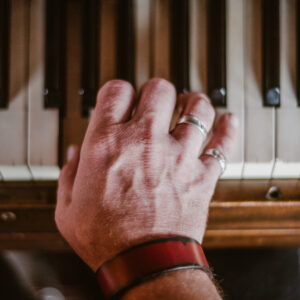 Une main portant des bagues en argent et un bracelet en cuir rouge repose sur les touches d'un piano ancien.