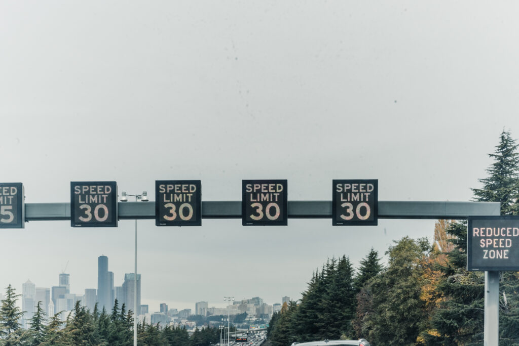 Des panneaux lumineux affichant une limite de vitesse de 30 surplombent une autoroute bordée d'arbres, avec une ville visible au loin.