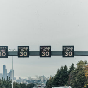 Des panneaux lumineux affichant une limite de vitesse de 30 surplombent une autoroute bordée d'arbres, avec une ville visible au loin.