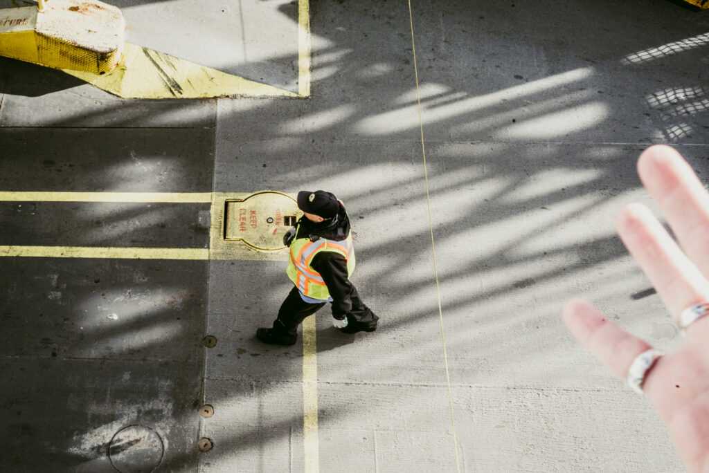 Vue en plongée d'un ouvrier en gilet de sécurité jaune marchant sur un sol en béton, avec une main tendue au premier plan.