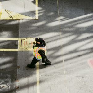 Vue en plongée d'un ouvrier en gilet de sécurité jaune marchant sur un sol en béton, avec une main tendue au premier plan.