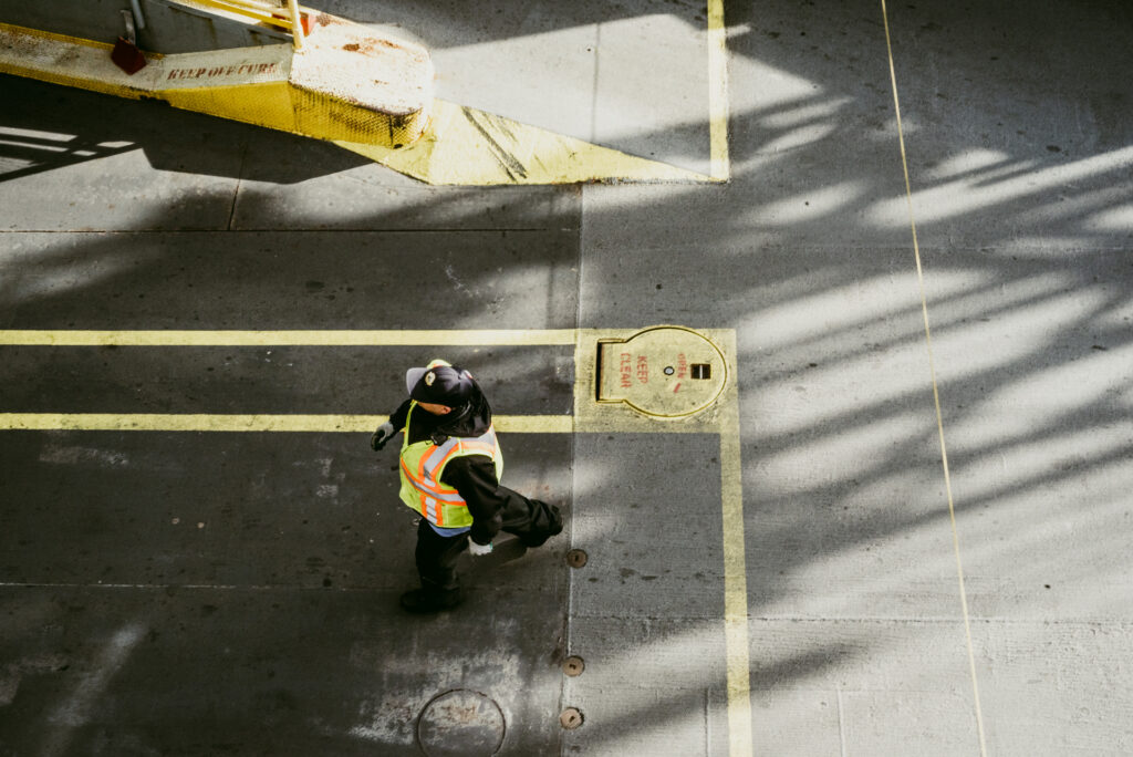 Vue en plongée d'une personne en gilet de sécurité fluorescent marchant sur un sol en béton marqué de lignes jaunes, avec de fortes ombres projetées.