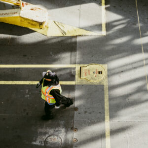 Vue en plongée d'une personne en gilet de sécurité fluorescent marchant sur un sol en béton marqué de lignes jaunes, avec de fortes ombres projetées.