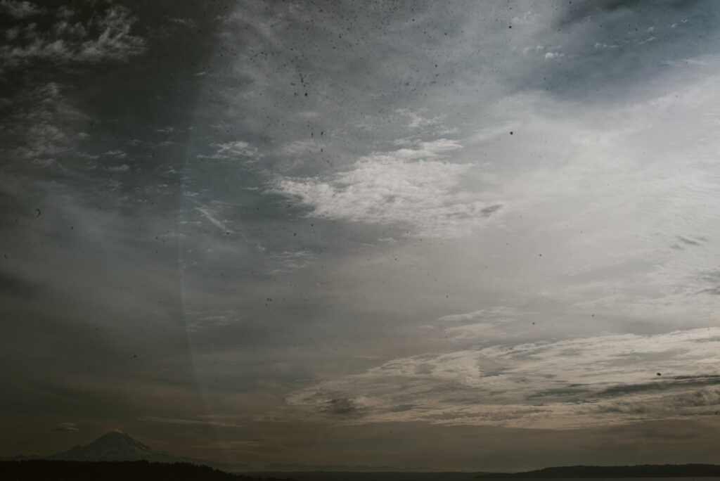 Un ciel nuageux et lumineux surplombe une montagne enneigée à l'horizon, avec des collines boisées en avant-plan.