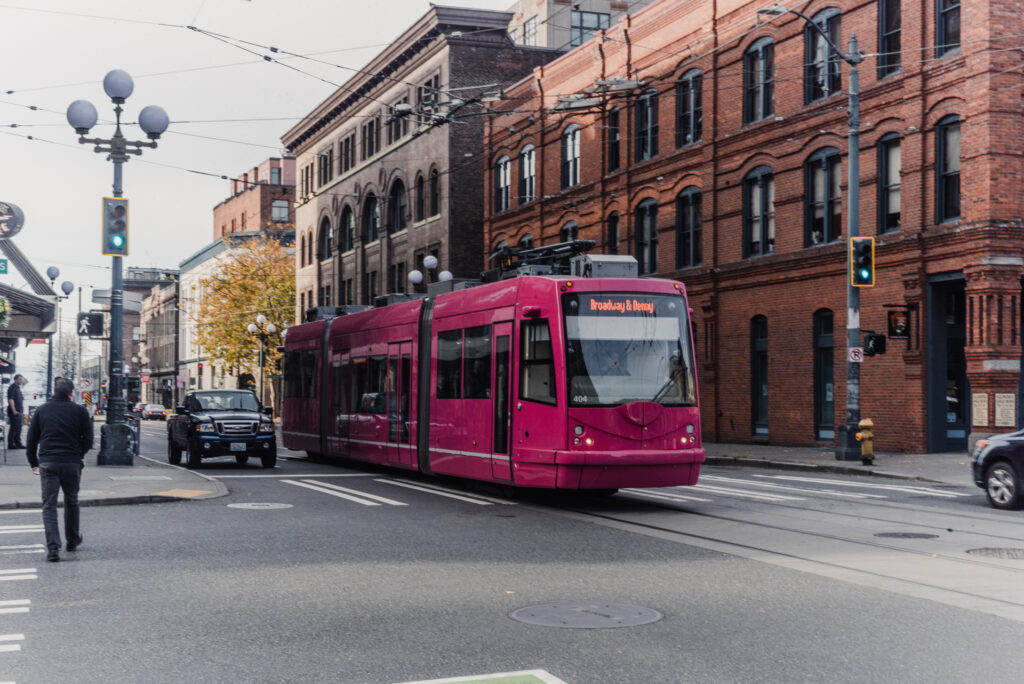 Un tramway rose circule dans une rue bordée de bâtiments en briques rouges, tandis qu'un piéton traverse au passage protégé.