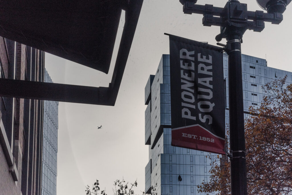 Une bannière verticale indiquant 'Pioneer Square, Est. 1852' est fixée à un lampadaire devant des immeubles en verre, avec un avion visible dans le ciel gris.